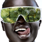 Close-up of a smiling person wearing clear wraparound goggles filled with broccoli florets, with their tongue touching the corner of their mouth.