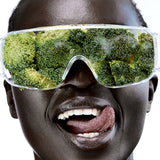 Close-up of a smiling person wearing clear wraparound goggles filled with broccoli florets, with their tongue touching the corner of their mouth.