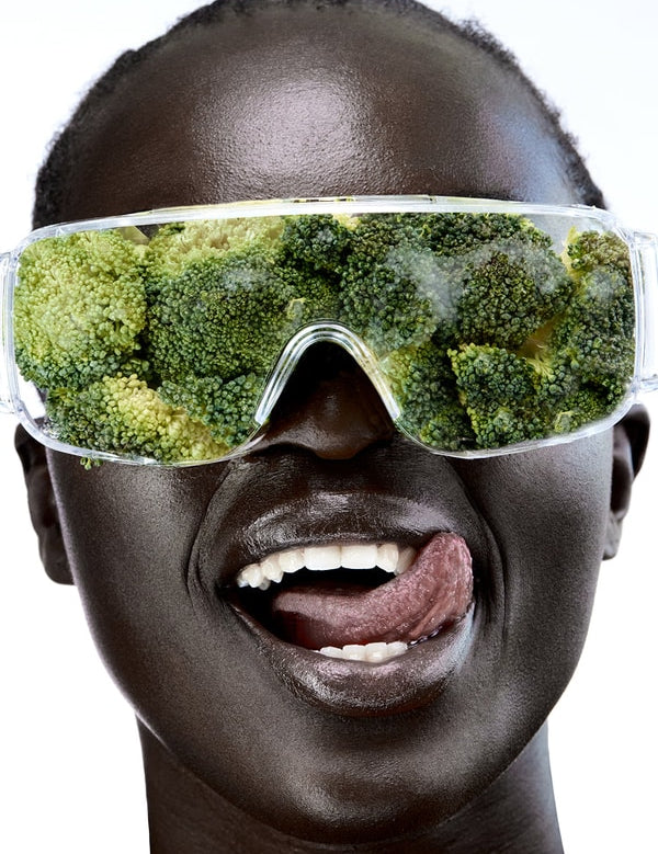 Close-up of a smiling person wearing clear wraparound goggles filled with broccoli florets, with their tongue touching the corner of their mouth.