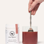 A hand stirring a reddish-brown drink in a clear glass with a metal spoon beside a white The Beauty Chef Glow Ageless jar with a pink strip.