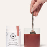 A hand stirring a reddish-brown drink in a clear glass with a metal spoon beside a white The Beauty Chef Glow Ageless jar with a pink strip.