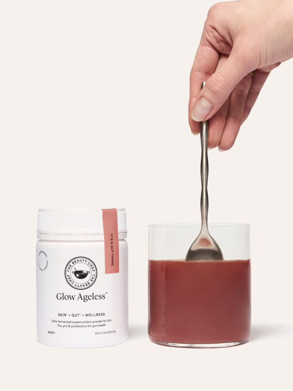 A hand stirring a reddish-brown drink in a clear glass with a metal spoon beside a white The Beauty Chef Glow Ageless jar with a pink strip.