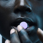 Close-up of a person holding a round pale-purple The Beauty Chef Plumpers Kakadu Plum and Berry tablet at their parted lips, with cool blue lighting across their face and fingers.