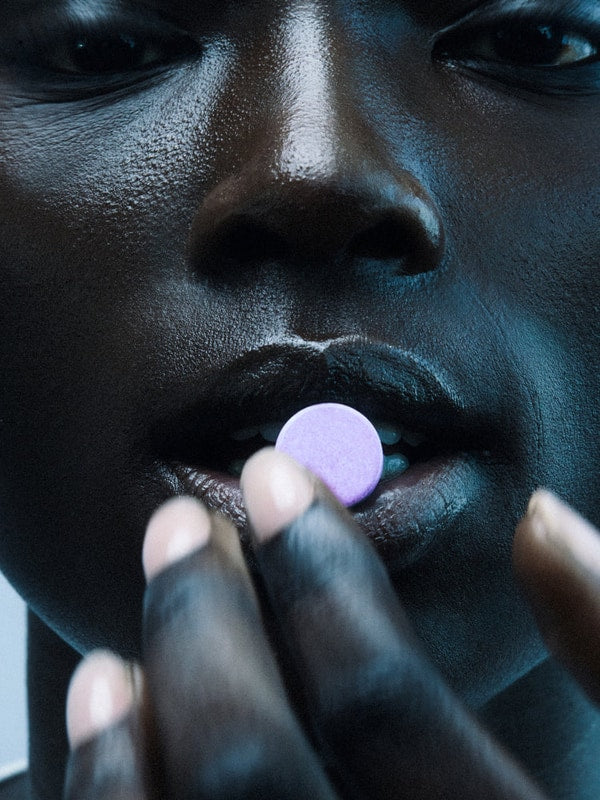 Close-up of a person holding a round pale-purple The Beauty Chef Plumpers Kakadu Plum and Berry tablet at their parted lips, with cool blue lighting across their face and fingers.