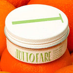 A white jar labeled “Tuttofare Sicilian Citrus Tallow Balm 70g” with green text and a metal lid marked by a green line sits atop bright oranges, covered in droplets. This hydrating balm is crafted from grass-fed beef tallow.