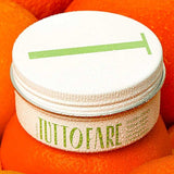 A white jar labeled “Tuttofare Sicilian Citrus Tallow Balm 70g” with green text and a metal lid marked by a green line sits atop bright oranges, covered in droplets. This hydrating balm is crafted from grass-fed beef tallow.