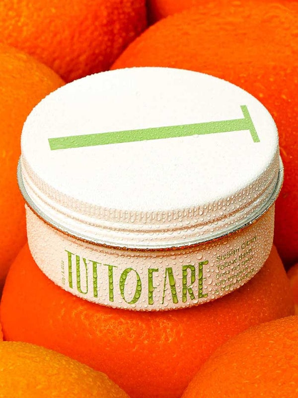A white jar labeled “Tuttofare Sicilian Citrus Tallow Balm 70g” with green text and a metal lid marked by a green line sits atop bright oranges, covered in droplets. This hydrating balm is crafted from grass-fed beef tallow.