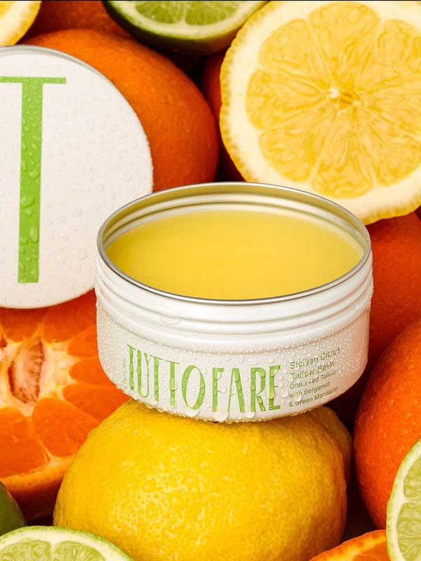 An open tin of Tuttofare Sicilian Citrus Tallow Balm 70g, made with grass-fed beef tallow, sits among sliced lemons, limes, and oranges. Its hydrating yellow balm and dewy tin capture the essence of pure citrus freshness.