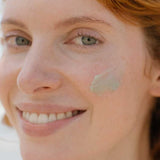 Close-up of a smiling woman with fair skin and auburn hair, gently applying a small amount of Tuttofare Roman Blue Tallow Balm 70g to her right cheek.