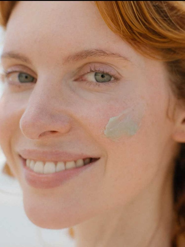 Close-up of a smiling woman with fair skin and auburn hair, gently applying a small amount of Tuttofare Roman Blue Tallow Balm 70g to her right cheek.