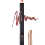 A tan nude INIKA Organic Lip Crayons 6.7g stands upright with its cap off, tip exposed. Behind it is a swatch of the long-lasting liner and "INIKA Organic Lip Crayon" with "Tan Nude" below on the product.
