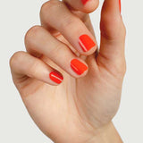 A close-up of a hand with neatly manicured nails painted in Sienna Byron Bay Nail Polish - Tango, a glossy tangerine orange shade, against a plain light background.