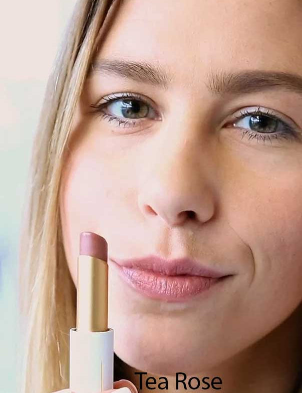 A woman with light skin applies Luk Beautifood Lip Nourish Lipsticks 3g in Tea Rose, holding the mauve-pink tube near her face. The words "Tea Rose" appear at the bottom of the image.