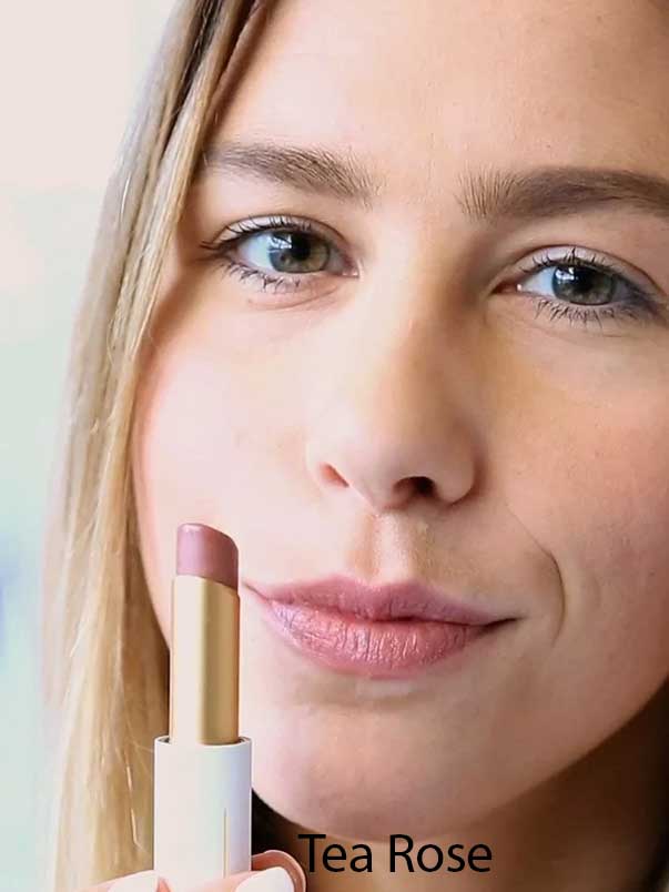 A woman with light skin applies Luk Beautifood Lip Nourish Lipsticks 3g in Tea Rose, holding the mauve-pink tube near her face. The words "Tea Rose" appear at the bottom of the image.