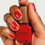 A hand with red painted nails holds a bottle of Sienna Byron Bay Nail Polish - Tempest, featuring a wooden cap. The background is plain white.