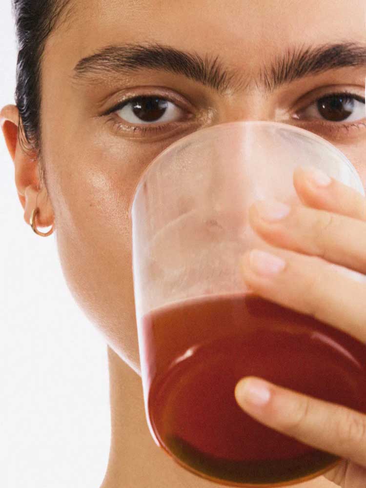 A person with a small gold hoop earring holds a clear glass of reddish-brown liquid—possibly The Beauty Chef Clear Skin™ Berry 150g—close to their mouth, partially obscuring their clear skin against a plain white background.