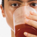 A person with a small gold hoop earring holds a clear glass of reddish-brown liquid—possibly The Beauty Chef Clear Skin™ Berry 150g—close to their mouth, partially obscuring their clear skin against a plain white background.