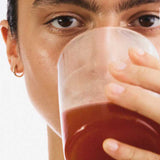 A person with a small gold hoop earring holds a clear glass of reddish-brown liquid—possibly The Beauty Chef Clear Skin™ Berry 150g—close to their mouth, partially obscuring their clear skin against a plain white background.