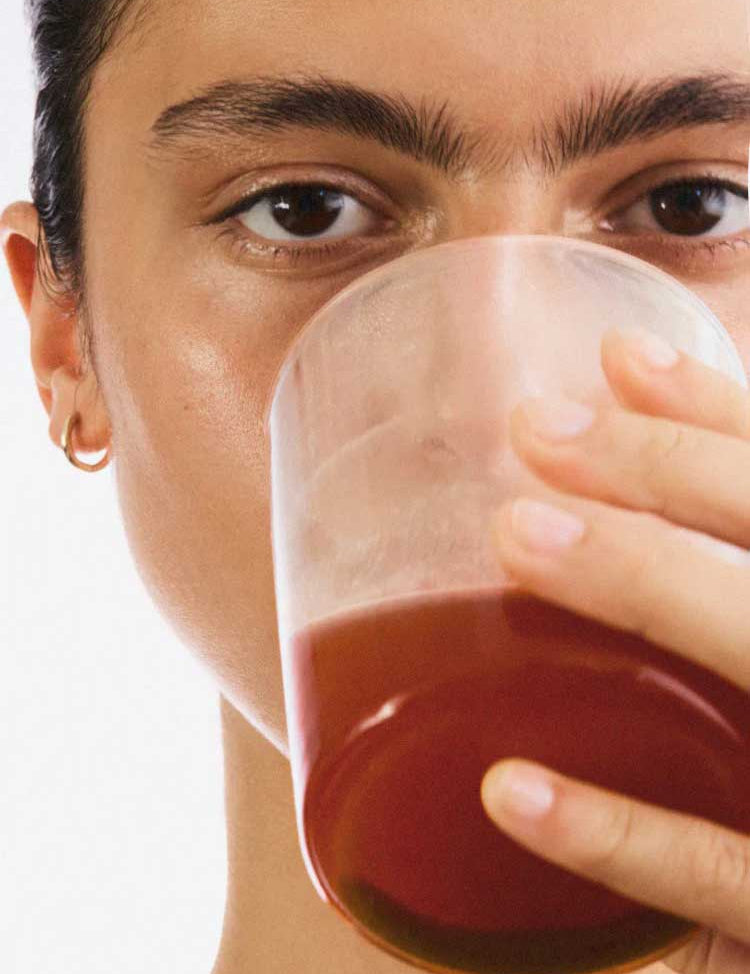 A person with a small gold hoop earring holds a clear glass of reddish-brown liquid—possibly The Beauty Chef Clear Skin™ Berry 150g—close to their mouth, partially obscuring their clear skin against a plain white background.