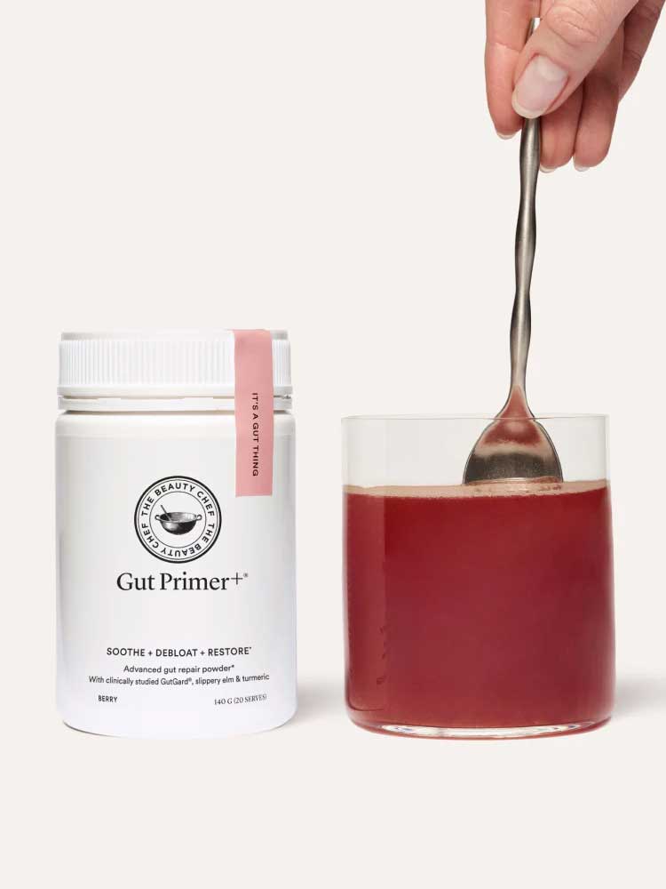 A container of The Beauty Chef GUT PRIMER+® 140g gut health powder stands next to a glass of red liquid, being stirred by hand against a plain, light background, highlighting GutGard benefits and relief from abdominal bloating.