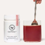 A container of The Beauty Chef GUT PRIMER+® 140g gut health powder stands next to a glass of red liquid, being stirred by hand against a plain, light background, highlighting GutGard benefits and relief from abdominal bloating.