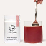 A container of The Beauty Chef GUT PRIMER+® 140g gut health powder stands next to a glass of red liquid, being stirred by hand against a plain, light background, highlighting GutGard benefits and relief from abdominal bloating.