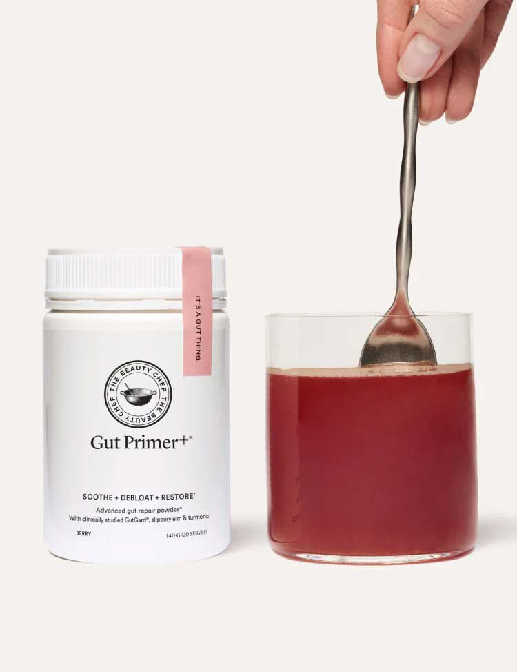 A container of The Beauty Chef GUT PRIMER+® 140g gut health powder stands next to a glass of red liquid, being stirred by hand against a plain, light background, highlighting GutGard benefits and relief from abdominal bloating.