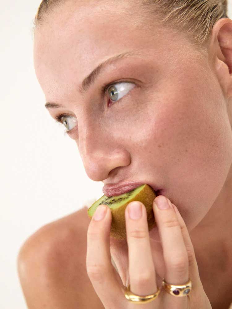 Close-up of a light-skinned, blue-eyed person with gold rings holding and about to bite into a half-peeled kiwi—a natural prebiotic fiber source—promoting gut health with The Beauty Chef The Perfect Poo™ 270g. Light, plain background.
