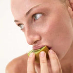 Close-up of a light-skinned, blue-eyed person with gold rings holding and about to bite into a half-peeled kiwi—a natural prebiotic fiber source—promoting gut health with The Beauty Chef The Perfect Poo™ 270g. Light, plain background.