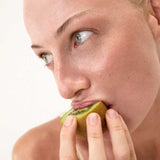 Close-up of a light-skinned, blue-eyed person with gold rings holding and about to bite into a half-peeled kiwi—a natural prebiotic fiber source—promoting gut health with The Beauty Chef The Perfect Poo™ 270g. Light, plain background.