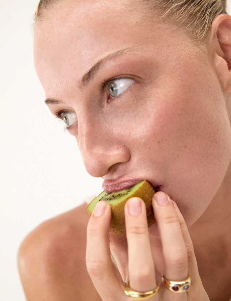 Close-up of a light-skinned, blue-eyed person with gold rings holding and about to bite into a half-peeled kiwi, a natural prebiotic fiber source, promoting gut health with The Beauty Chef The Perfect Poo™ 270g. Light, plain background.