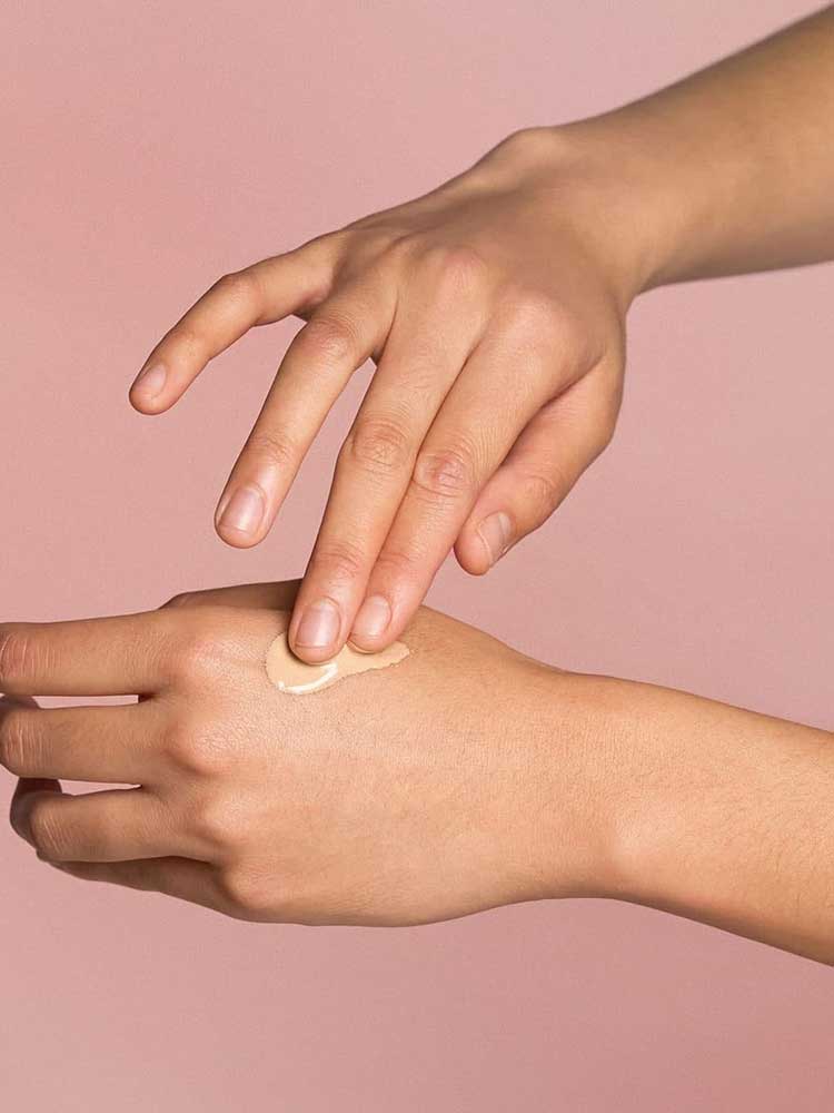 A person applies The Jojoba Company Illuminating Tinted Moisturiser SPF15 50ml to the back of their hand with their fingers, set against a soft pink background.