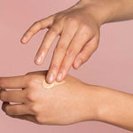 A person applies The Jojoba Company Illuminating Tinted Moisturiser SPF15 50ml to the back of their hand with their fingers, set against a soft pink background.