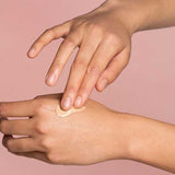 A person applies The Jojoba Company Illuminating Tinted Moisturiser SPF15 50ml to the back of their hand with their fingers, set against a soft pink background.