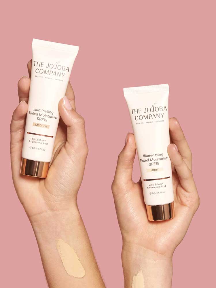 Two hands hold tubes of The Jojoba Company Illuminating Tinted Moisturiser SPF15 50ml against a pink background, with swatches on each hand showing the diverse shades offered.