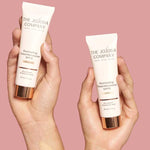 Two hands hold tubes of The Jojoba Company Illuminating Tinted Moisturiser SPF15 50ml against a pink background, with swatches on each hand showing the diverse shades offered.