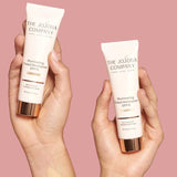 Two hands hold tubes of The Jojoba Company Illuminating Tinted Moisturiser SPF15 50ml against a pink background, with swatches on each hand showing the diverse shades offered.