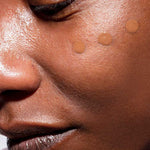 Close-up of a person’s face with three dots of The Organic Skin Co The Only Glow Tinted Face Oil - Deep 30ml on their cheek, highlighting a luminous complexion and smooth makeup application.
