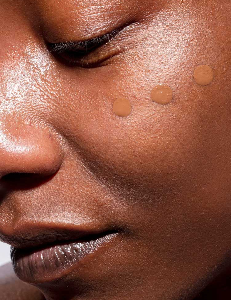 Close-up of a person’s face with three dots of The Organic Skin Co The Only Glow Tinted Face Oil - Deep 30ml on their cheek, highlighting a luminous complexion and smooth makeup application.
