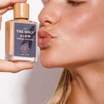 A woman with glowing skin holds The Organic Skin Co The Only Glow Tinted Face Oil - Light 30ml near her face, pursing her lips as if to kiss the bottle, which has a wooden cap and minimalist label, promising a luminous dewy finish.