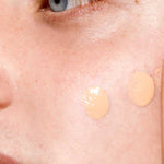 Close-up of a person's face with two beige foundation dots, showcasing smooth skin and a blue eye—an ideal base for The Organic Skin Co The Only Glow Tinted Face Oil - Light 30ml to achieve a luminous, dewy finish.