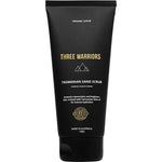 A black tube labeled Three Warriors Tasmanian Sand Scrub 150g features gold and white text, a mountain logo, and highlights its use as an organic exfoliant for intense hydration and preparing skin for self-tanning.