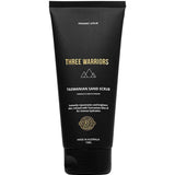 A black tube labeled Three Warriors Tasmanian Sand Scrub 150g features gold and white text, a mountain logo, and highlights its use as an organic exfoliant for intense hydration and preparing skin for self-tanning.