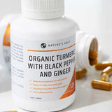 A bottle of Nature's Help Turmeric Capsules with Black Pepper & Ginger sits on a white surface, surrounded by turmeric capsules and a blurred bottle in the background. The label highlights health benefits and contains 120 capsules.