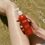 A person’s legs are submerged in clear, shallow water as they hold a red bottle of Tuttofare Not Your Regular Body Oil 200ml. Sunlight reflects off their skin and the bottle, evoking a fresh, summery vibe.