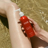 A person’s legs are submerged in clear, shallow water as they hold a red bottle of Tuttofare Not Your Regular Body Oil 200ml. Sunlight reflects off their skin and the bottle, evoking a fresh, summery vibe.
