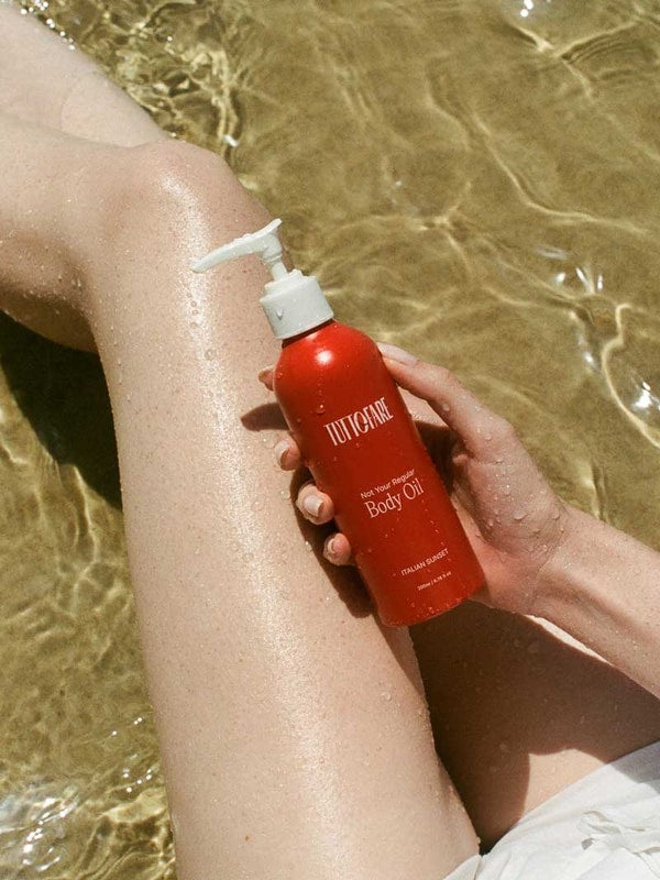 A person’s legs are submerged in clear, shallow water as they hold a red bottle of Tuttofare Not Your Regular Body Oil 200ml. Sunlight reflects off their skin and the bottle, evoking a fresh, summery vibe.