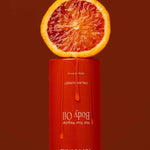 A halved blood orange sits on an upside-down Tuttofare Not Your Regular Body Oil 200ml bottle with orange liquid dripping down, set against a deep red background, emphasizing this luxurious natural moisturizer.