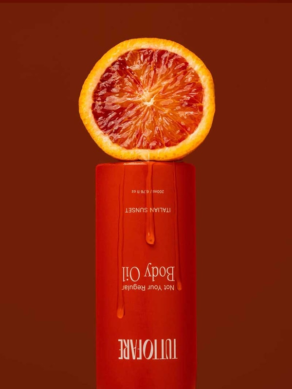 A halved blood orange sits on an upside-down Tuttofare Not Your Regular Body Oil 200ml bottle with orange liquid dripping down, set against a deep red background, emphasizing this luxurious natural moisturizer.