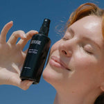 A person with fair skin and reddish hair smiles with closed eyes, holding a black bottle labeled Tuttofare Naturally Active Face Mist 100ml near their face against a blue sky background.